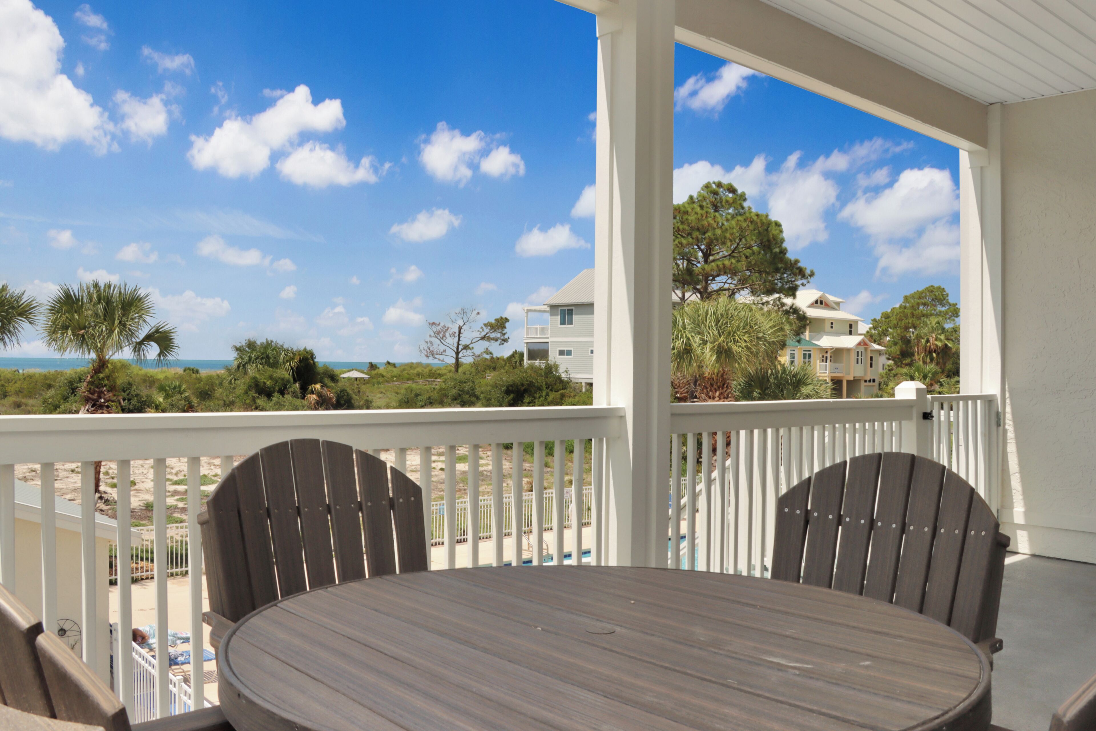 Eine überdachte Terrasse im Freien mit einem runden Tisch und zwei Stühlen, mit Blick auf einen Strand und ein Wohngebiet.