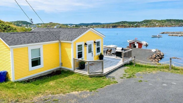 A yellow house with a porch, a small deck, and a view of the water and hills in the background. - Carriage Hill Cottage (Twillingate)