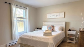 A neatly arranged bedroom with a bed, bedside tables, a nightstand, a wall-mounted sign, and a window with curtains. - Carriage Hill Cottage (Twillingate)