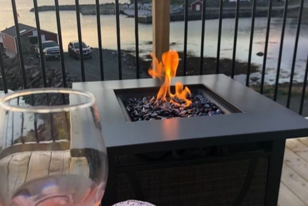 Ett glasbord med ett vinglas på, en eldstad med ved och en utsikt över en vattenyta och byggnader i bakgrunden.
