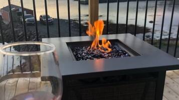 Una mesa de vidrio con una copa de vino, una chimenea con leña y una vista de agua y edificios al fondo.