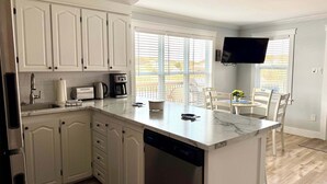 A modern kitchen with white cabinetry, a built-in dishwasher, a coffee maker, and a dining area with a table and chairs. - Carriage Hill Cottage (Twillingate)