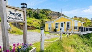 A wooden sign with a horse and carriage, a yellow building with a porch, and a fenced area with flowers. - Carriage Hill Cottage (Twillingate)