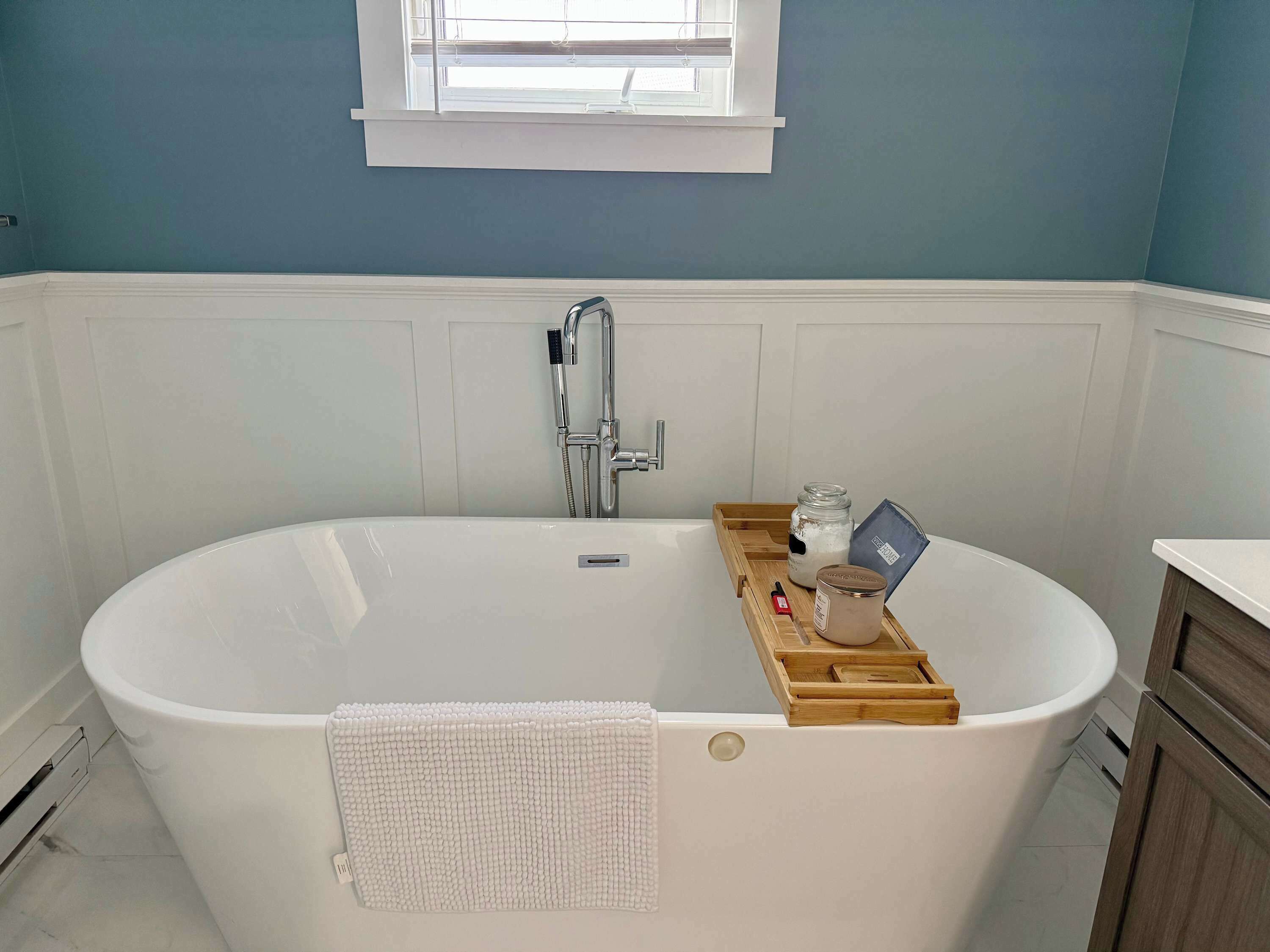 A modern bathroom with a freestanding bathtub, a window above the tub, and a wooden tray with toiletries.