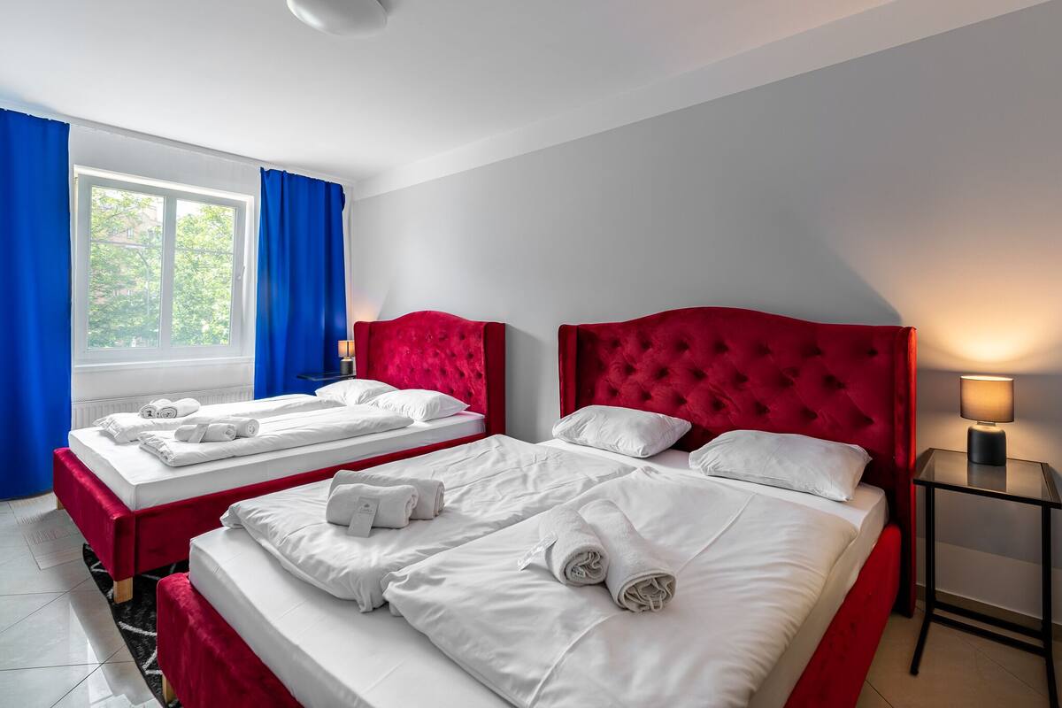Une chambre d’hôtel avec deux lits, des têtes de lit rouges, du linge de lit blanc, une fenêtre avec des rideaux bleus et une table de chevet avec une lampe.