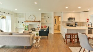 A modern kitchen with white cabinets, a microwave, and a coffee maker. There is a dining area with a table and chairs, and a living room with a sofa and a coffee table.