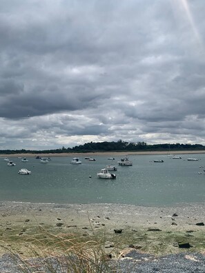 A coastal scene with boats on calm water, a sandy beach, and a forested area in the background.