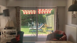 A living room with a sliding glass door leading to a patio with outdoor furniture and a striped awning. - Appartement 8 Personnes, 3mn de la mer , 1 mn de la Thalasso, Centre Ville, (Ouistreham)