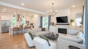 A spacious living room with a large sofa set, a coffee table, and a TV mounted on the wall. There is a kitchen area with a dining table and chairs, and a fireplace.