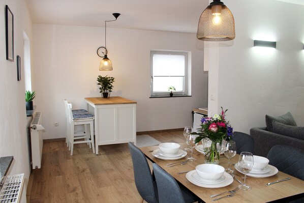 A dining area with a wooden table set for four, white chairs, a small kitchen area with a white countertop, and a window with blinds.