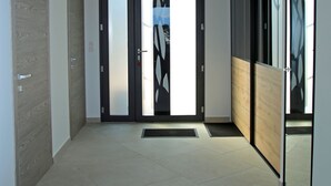 Ein moderner Flur mit einer Holztür, einer Glastür mit geometrischem Muster und einem großen Spiegel, der einen Teil des Raumes reflektiert.