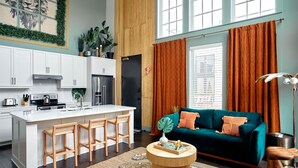 A modern kitchen with white cabinets, a central island with bar stools, and a dining area with a green sofa and orange curtains.