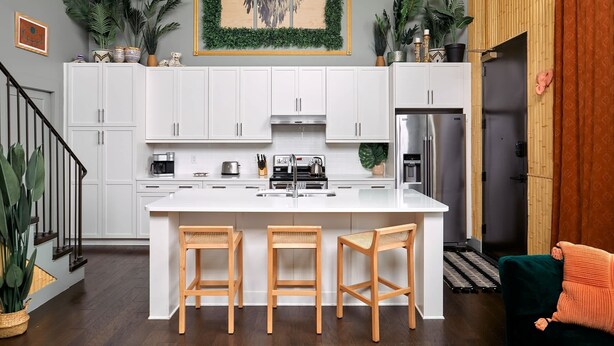 A modern kitchen with white cabinets, a central island with stools, and various plants.