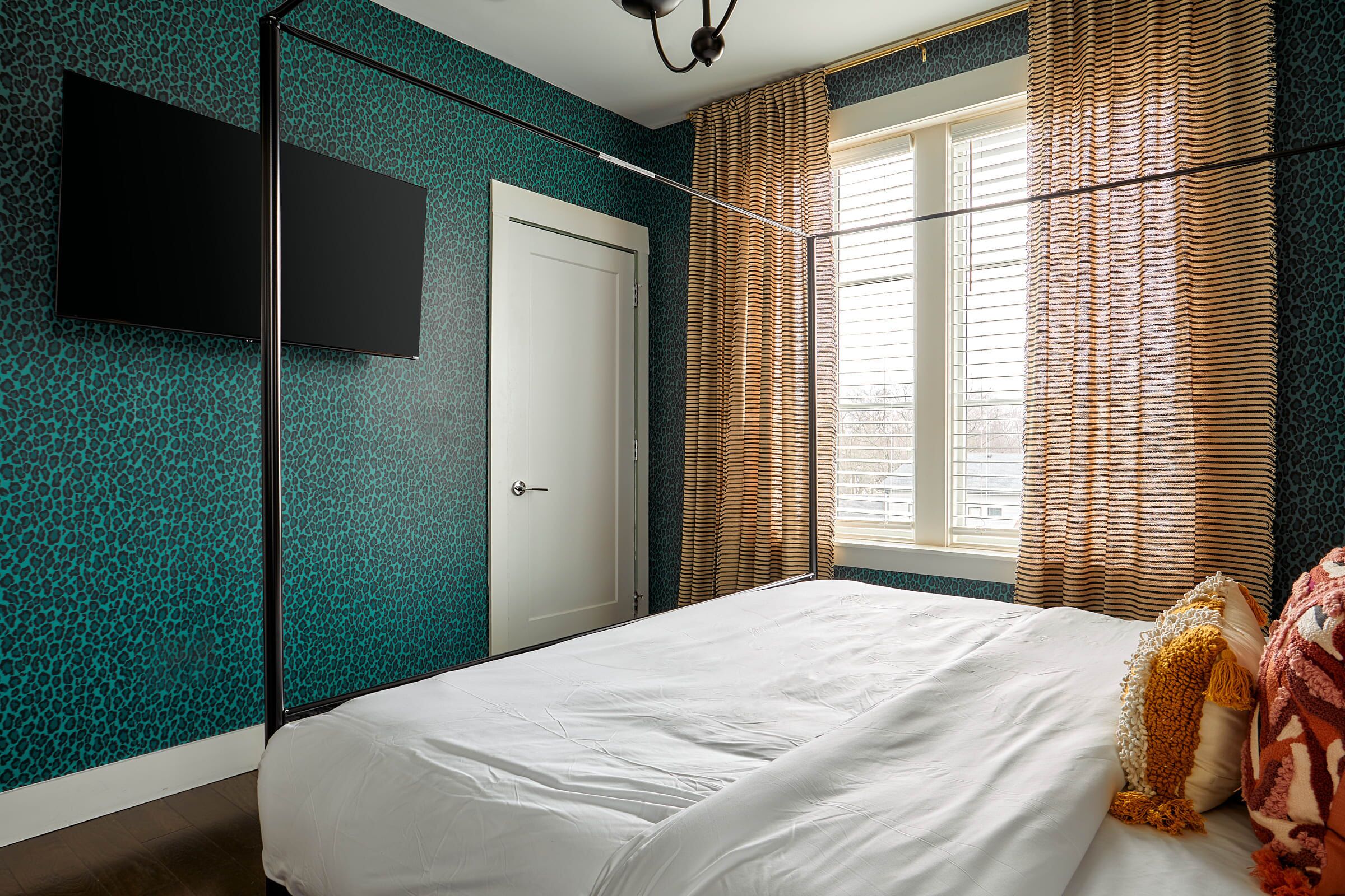 Ein Schlafzimmer mit einem Bett, einem an der Wand montierten Fernseher, einem Fenster mit Vorhängen und einer Tür.