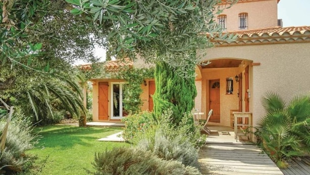 Una villa de estilo mediterráneo con terraza de madera, un porche cubierto y exuberante vegetación.