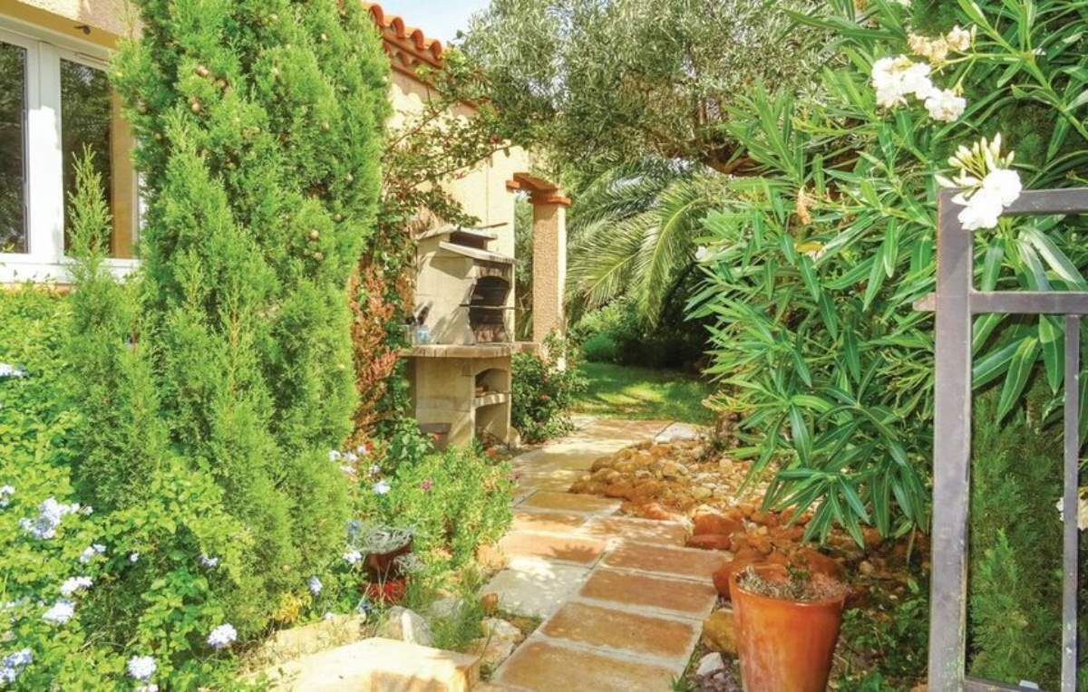 Un chemin de pierre mène à un espace de détente extérieur rustique, agrémenté d’une cheminée en pierre et entouré d’une végétation luxuriante et de plantes en fleurs.