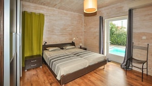 Un dormitorio con techo de madera, una cama con ropa de cama a rayas, una mesita de noche, una lámpara, una silla y vista a una piscina a través de una puerta de cristal.