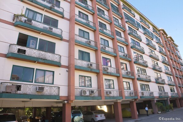 A multi-story hotel with balconies, air conditioning units, and a parking area with cars.