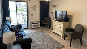 Ein Wohnzimmer mit einer Couch, einem Fernseher, einem Deckenventilator, einer Lampe, einem Sessel und Blick ins Freie.