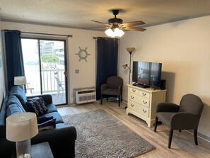 A living room with a sofa, a television, a ceiling fan, a lamp, a chair, and a view of the outside.