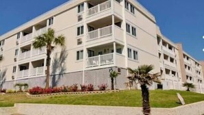 Ein mehrstöckiges Hotel mit Balkonen, einer flachen Dachterrasse und einer angelegten Vorderseite mit Palmen und Blumen.