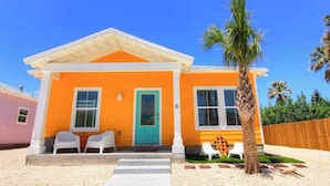 A two-story house with a bright orange facade, a turquoise door, and a small porch with white chairs. - Beach Please- Family Friendly | Community Pool (Port Aransas)
