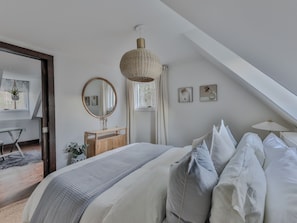Un dormitorio moderno con techo inclinado, una cama grande con ropa blanca, un aparador de madera, un espejo redondo y una lámpara colgante.