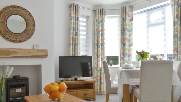 Una sala de estar moderna con un televisor de pantalla plana, muebles de madera, una mesa de comedor con sillas y ventanas amplias con cortinas.