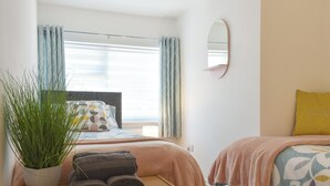Un dormitorio con cama, una mesita de noche con una planta y toallas, una ventana con cortinas y un espejo montado en la pared.