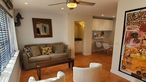 A living room with a sofa, two armchairs, a coffee table, a ceiling fan, a dining area with chairs, a kitchen with cabinets, and a large abstract painting.