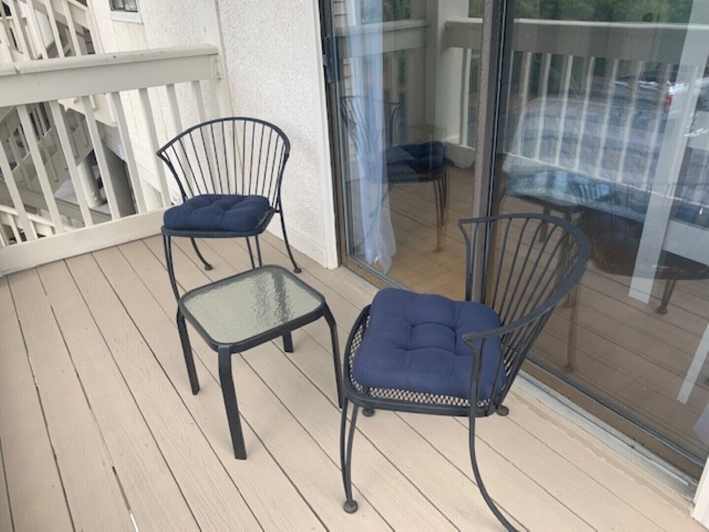 A balcony with two metal chairs and a small table, a white railing, and a glass door leading to a parking area.