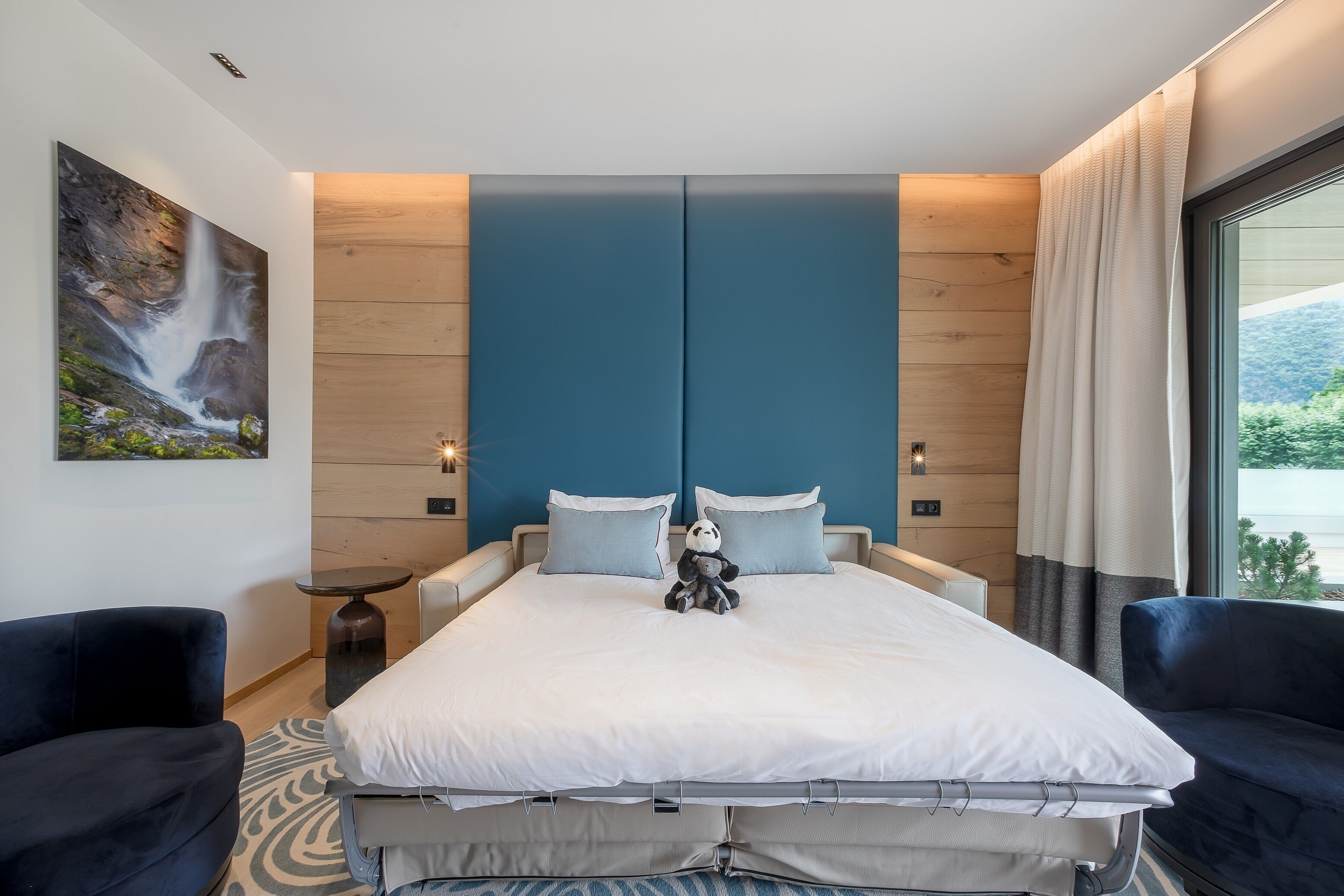 A modern bedroom with a large bed, two pillows, a nightstand, a small table, and a large window with curtains.