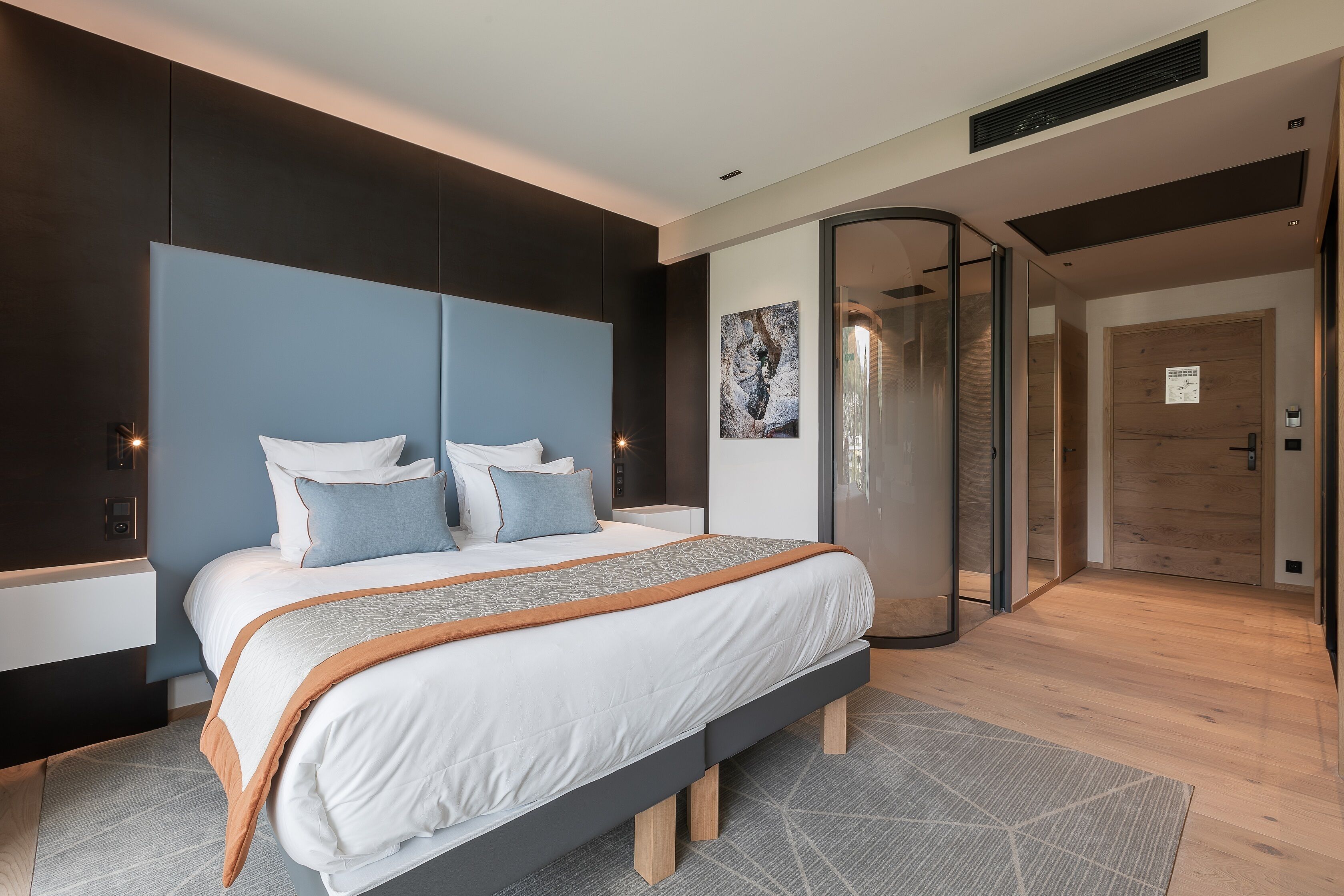 A modern bedroom with a large bed, a glass door leading to a bathroom, and a wooden floor.