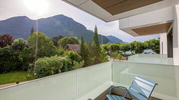 Un balcón con barandilla de vidrio, una banca de madera y vistas a montañas y vegetación.