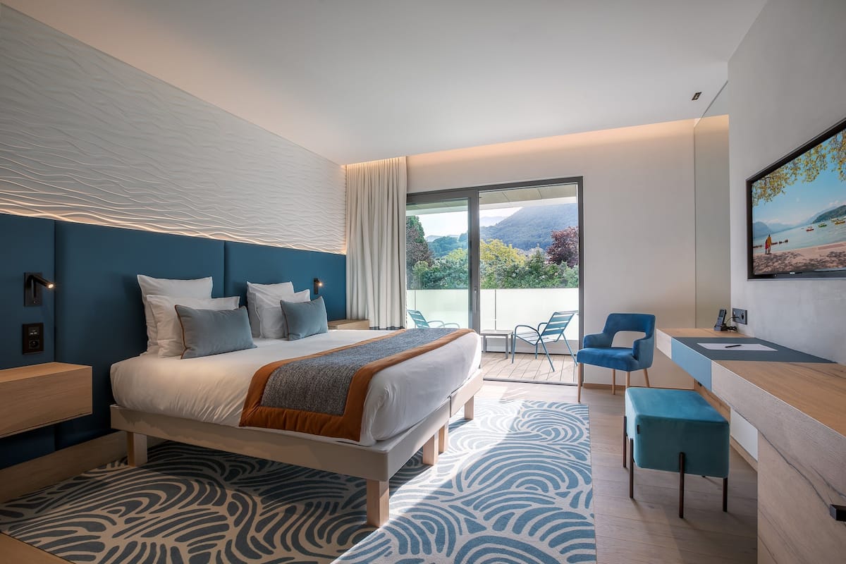 A modern hotel room with a large bed, a desk, and a balcony with a scenic view.