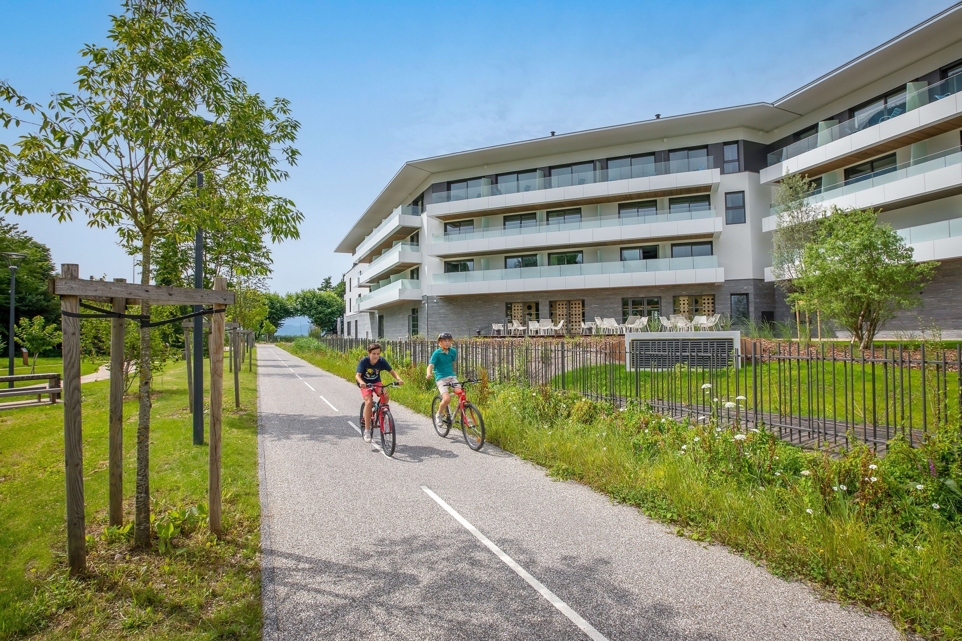 Een pad met twee fietsers, een modern gebouw met meerdere verdiepingen en een gazon met bomen.