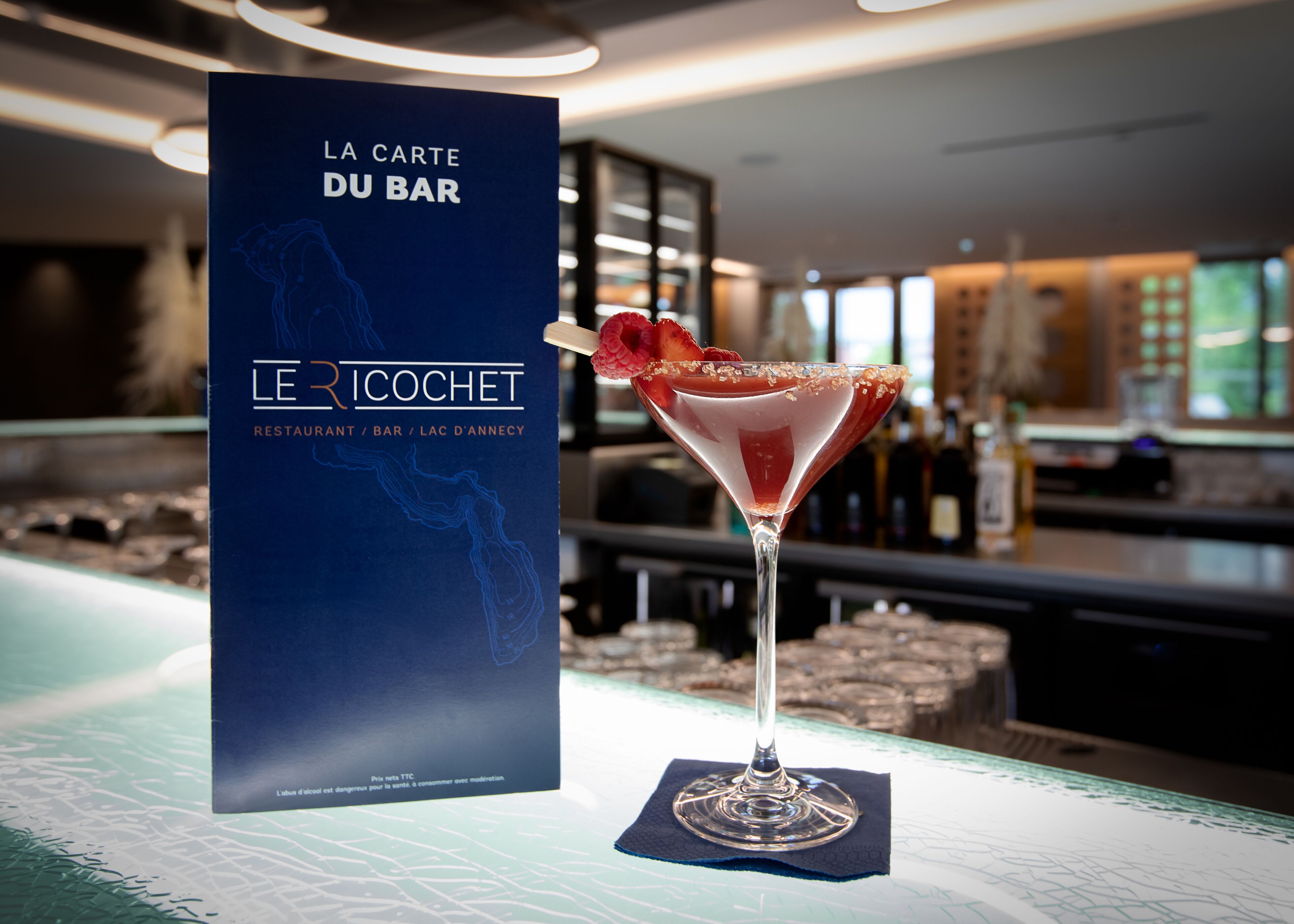 Un bicchiere da cocktail con una fragola sul bancone del bar, con un menu intitolato "LA CARTE DU BAR" sullo sfondo.