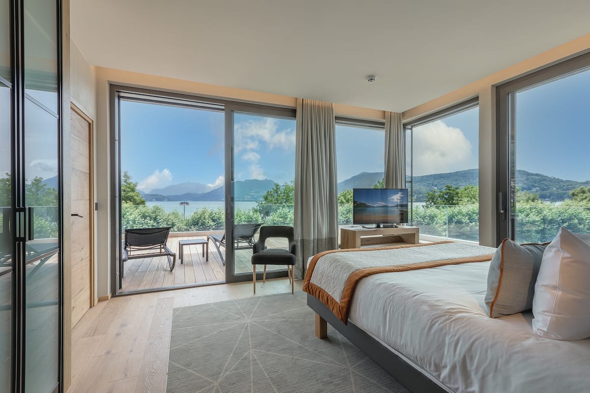 A modern bedroom with a large bed, a TV, and a balcony with a view of the mountains.