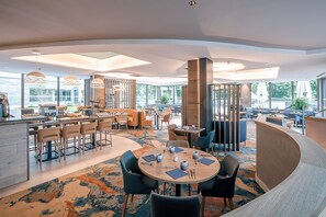 A modern hotel lobby with a central bar area, round tables with chairs, and large windows offering views of the outdoors.