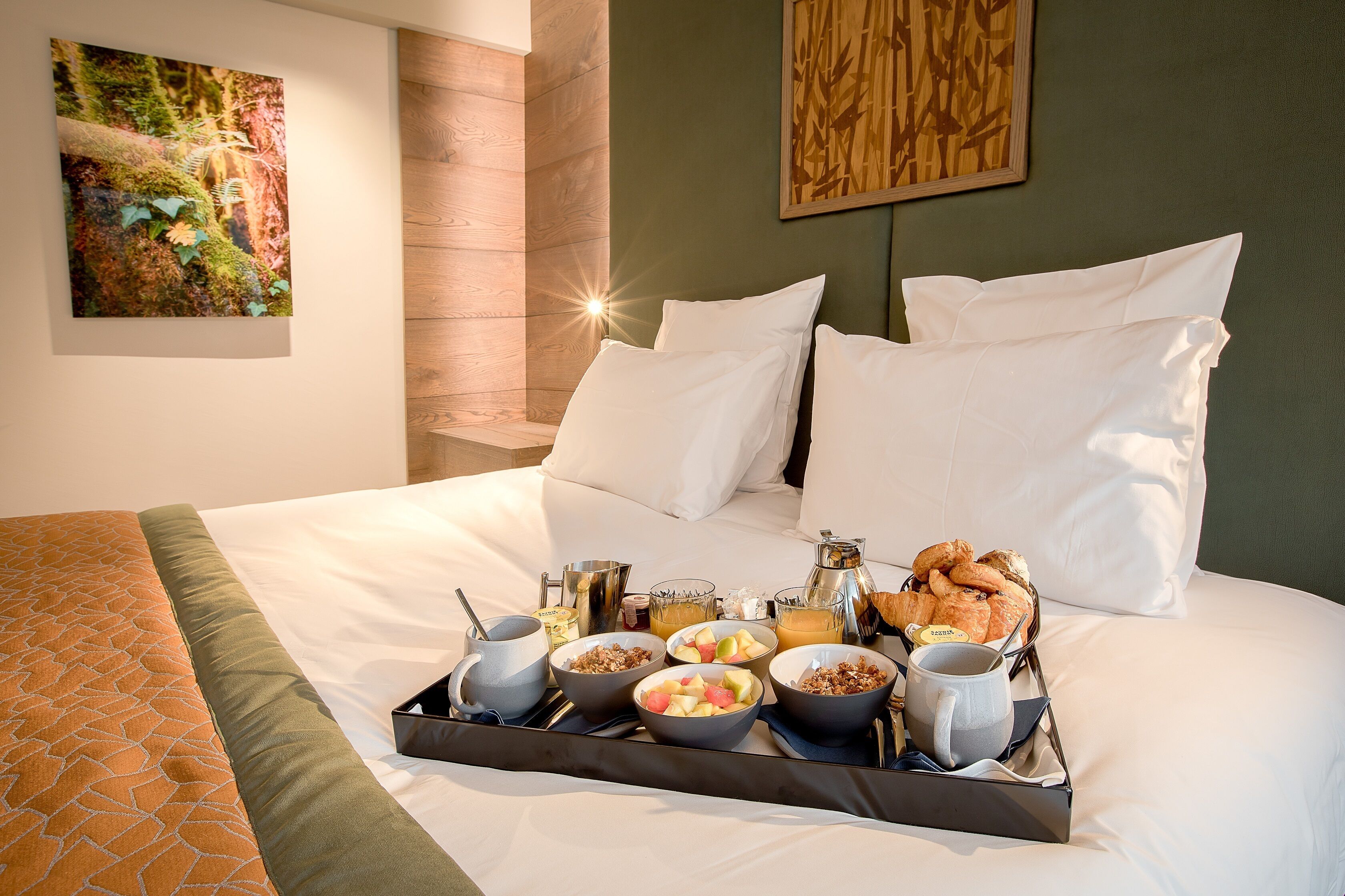 A neatly made bed with white linens, a tray with breakfast items including fruits, cereal, and pastries, and a painting on the wall.