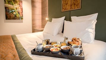 A neatly made bed with white linens, a tray with breakfast items including fruits, cereal, and pastries, and a painting on the wall.