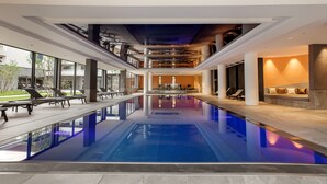Une piscine intérieure moderne avec des parois en verre transparent, un espace détente avec des sièges et une vue sur l’extérieur.