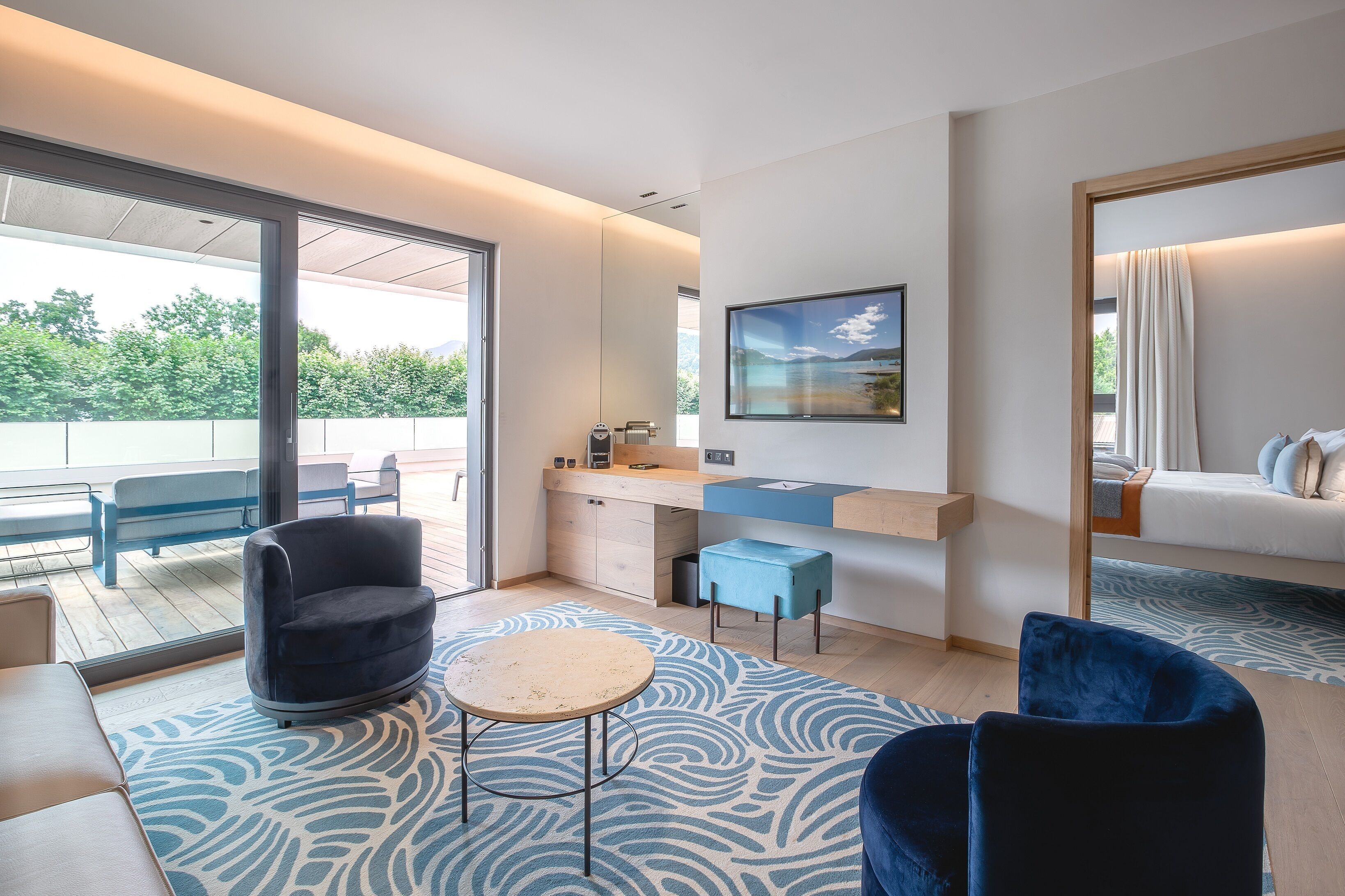 A modern hotel room with a large sliding glass door leading to a balcony, a small kitchen area, a seating area with a round table, and a large window with a view of greenery.