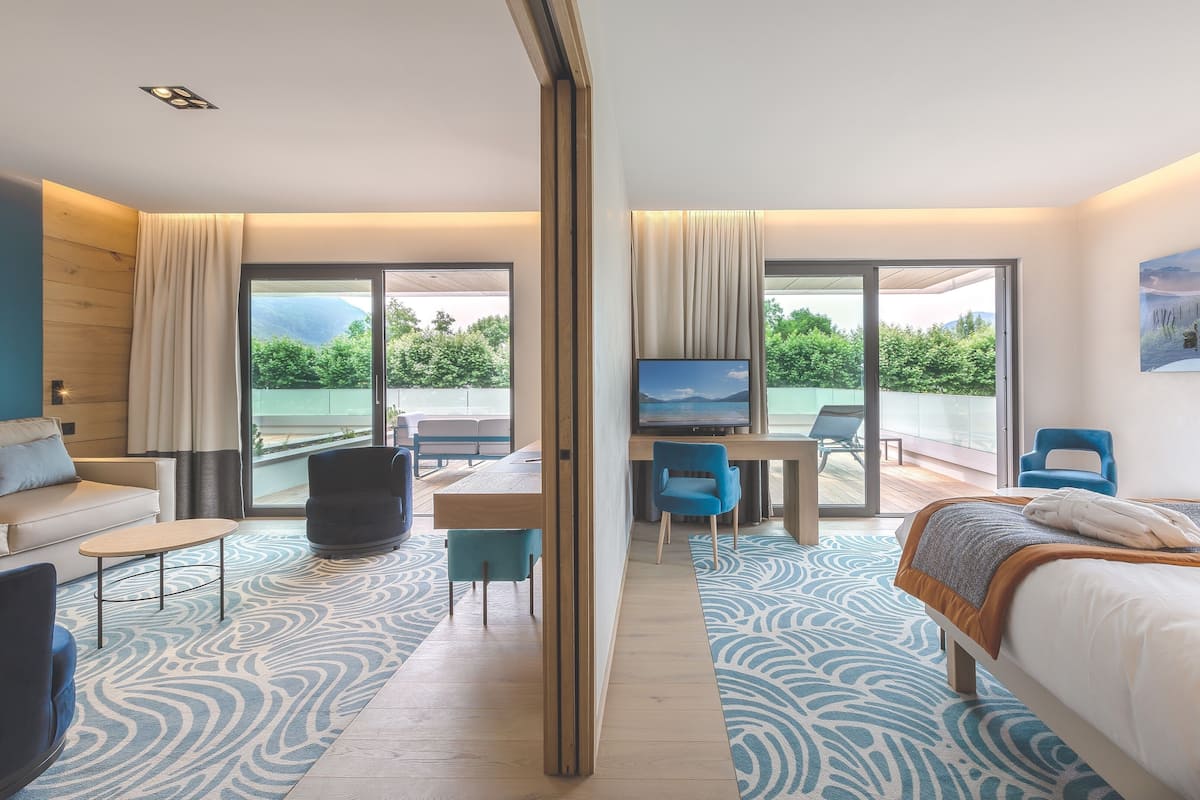 A modern hotel room with a large bed, a desk, a TV, and a balcony with a view of greenery.