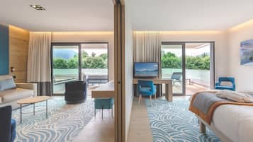 A modern hotel room with a large bed, a desk, a TV, and a balcony with a view of greenery.