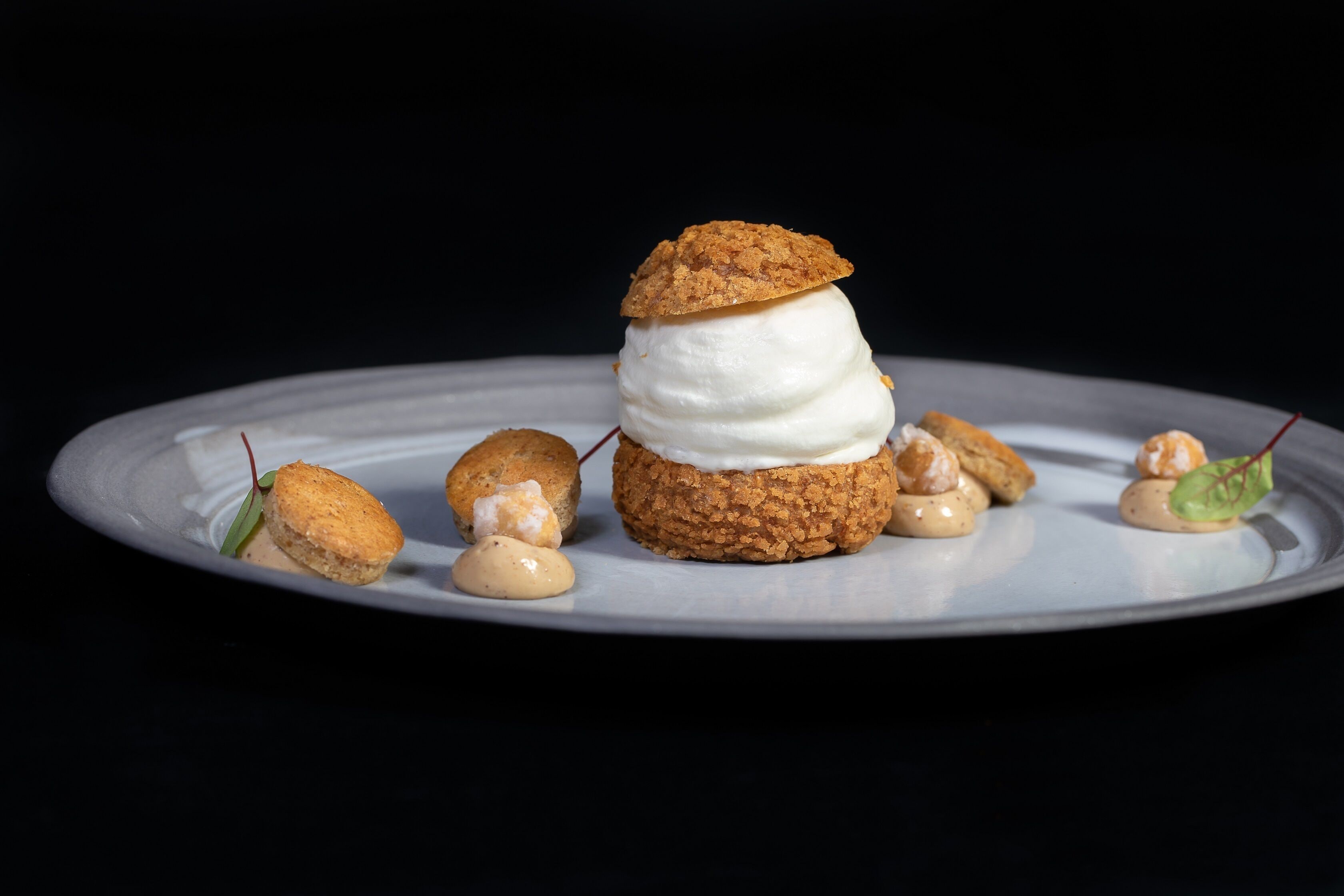 Un plato con un postre en capas, que incluye una galleta, malvaviscos y crema, acompañado de pequeñas cucharadas de crema y una decoración de hoja.