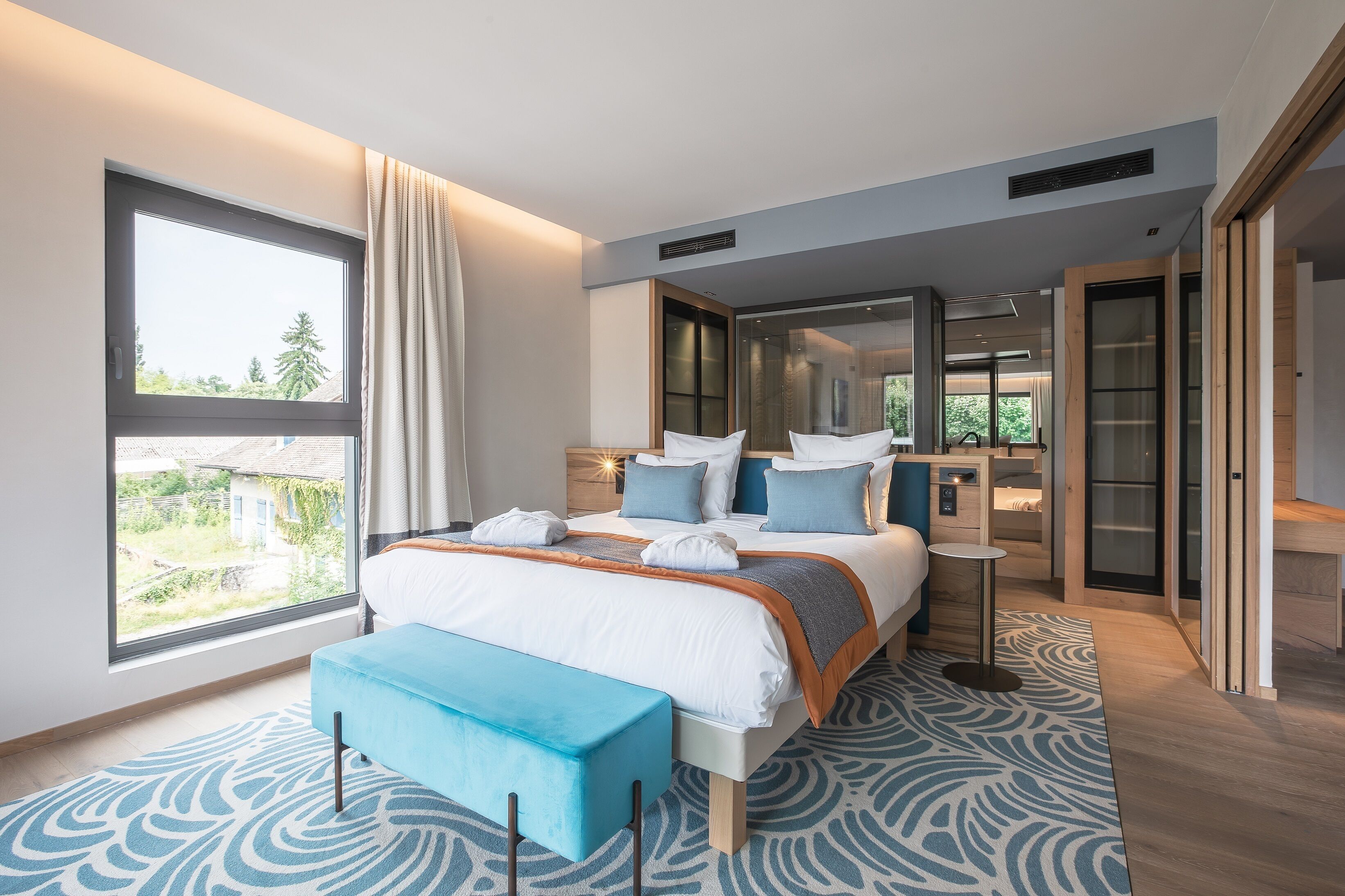A modern hotel room with a large bed, a view through a window, a bedside table, and a bench.