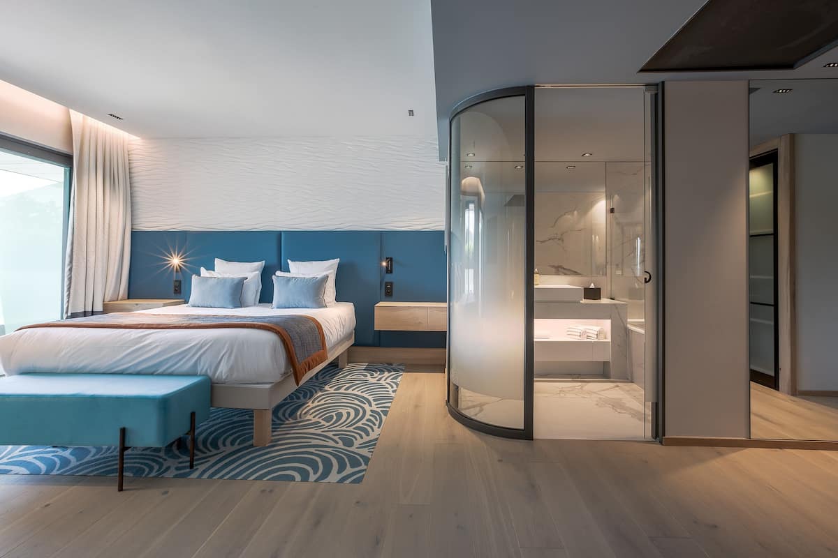 A modern bedroom with a bed, a bench, a glass-enclosed walk-in closet, and a bathroom with a marble floor.