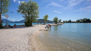 Une plage de galets bordée d’une eau claire, avec des arbres et des montagnes en arrière-plan.