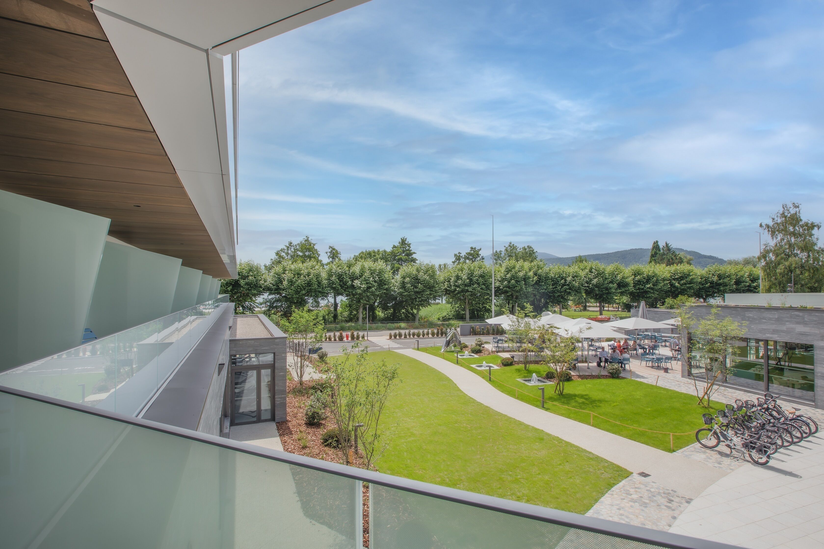 A modern building with a glass balcony overlooking a well-maintained courtyard with a grassy area, trees, and a bike parking area.